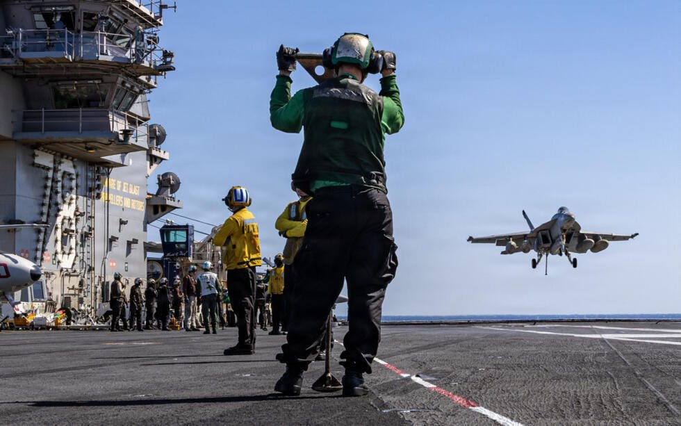 Cette photo fournie par la marine américaine montre des marins américains observant un avion F/A-18E Super Hornet s'approcher du pont d'envol de l'USS Gerald R. Ford lors de l'opération "Epic Fury",