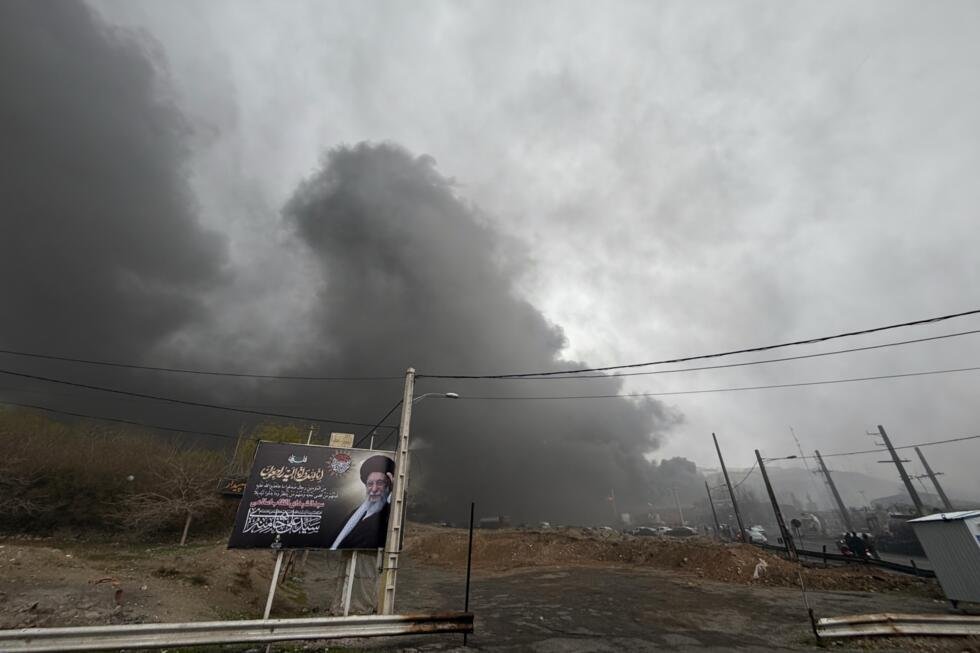 Une épaisse colonne de fumée persiste dans le ciel nuageux de Téhéran après une frappe contre une installation pétrolière, derrière un panneau d'affichage représentant le guide suprême Ali Khamenei.